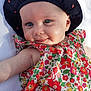 baby, infant, smiling, blue_eyes, hat, black_hat, apple_pattern, floral_dress, colorful_clothing, cute, lying_down, white_blanket, cheeky_smile, child, cute_face, skin, outdoor_light, portrait, baby_fashion, happy