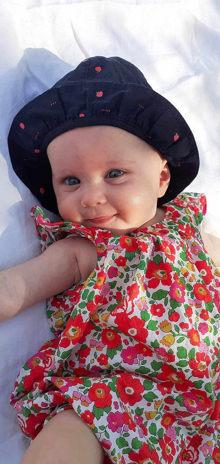 Prune a rejoint le concours — aidez-le/la à gagner de superbes lots ! baby, infant, smiling, blue_eyes, hat, black_hat, apple_pattern, floral_dress, colorful_clothing, cute, lying_down, white_blanket, cheeky_smile, child, cute_face, skin, outdoor_light, portrait, baby_fashion, happy