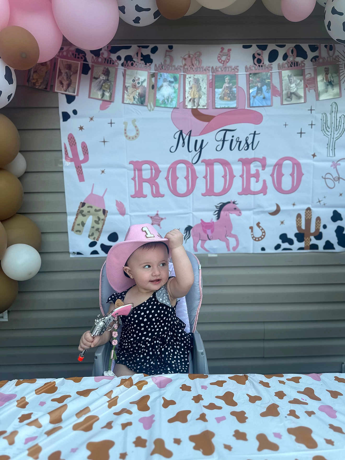 Aitana is registered to the contest to win money with this photo: baby, child, polka_dot, cowboy_hat, high_chair, toy_gun, balloons, party, tablecloth, cow_print, banner, pink, black, white, smiling, celebration, cute, indoor, portrait, festive