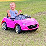 Aitana is registered to the contest to win money with this photo: toddler, child, toy_car, pink_car, mazda_mx5, grass, outdoor, happy, smiling, short_hair, casual_clothing, play, sunlight, daylight, greenery, small_vehicle, cute, joy, waving, person