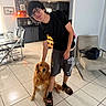 Athena joined the competition — help win amazing prizes! young_man, glasses, black_tshirt, shorts, slippers, golden_retriever, dog, indoor, kitchen, dining_area, tiled_floor, smiling, pet, chair, table, backpack, modern_interior, casual, home, portrait