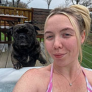 Archie is registered to the contest to win money with this photo: woman, dog, hot_tub, swimsuit, outdoor, deck, picnic_table, wood, fence, trees, cloudy_sky, smile, necklace, pet, water, relaxation, animal, portrait, canine, leisure