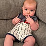 baby, infant, child, couch, sofa, clothing, striped_onesie, fingers_in_mouth, curious_expression, indoor, soft_lighting, plush_furniture, seated, barefoot, skin, head, face, person, cute, young