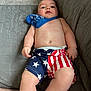 baby, child, infant, couch, sofa, bib, shorts, stars_and_stripes, pattern, clothing, indoor, furniture, relaxing, skin, face, legs, hand, portrait, casual, comfortable