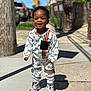 toddler, child, smiling, standing, sidewalk, outdoor, sunny, street, clothing, sneakers, black_hair, brick_wall, utility_pole, shadow, casual, urban, happy, portrait, daylight, person