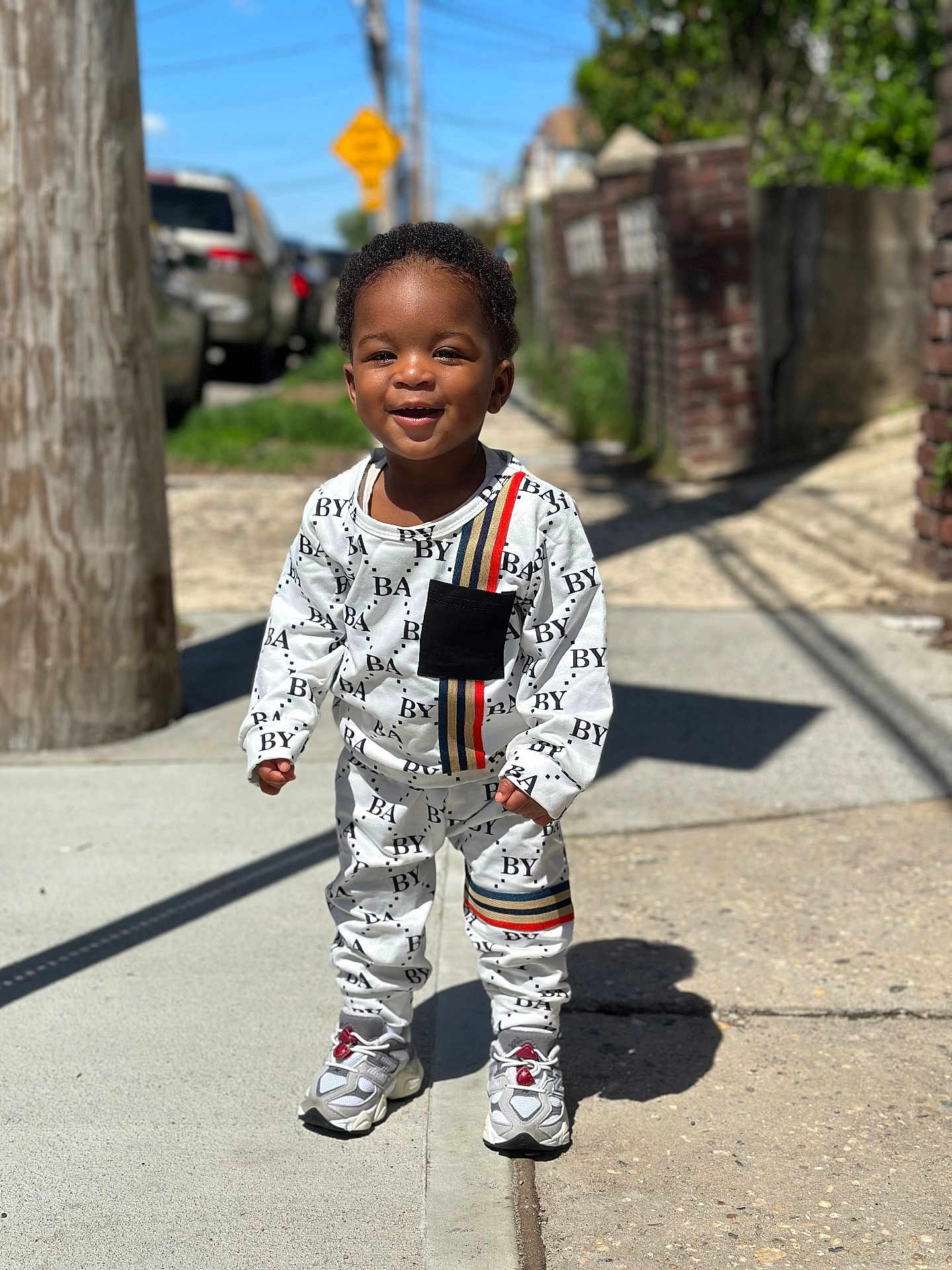 Kairo joined the competition — help win amazing prizes! toddler, child, smiling, standing, sidewalk, outdoor, sunny, street, clothing, sneakers, black_hair, brick_wall, utility_pole, shadow, casual, urban, happy, portrait, daylight, person