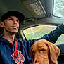 man, dog, car_interior, hoodie, cap, brown_dog, window, seat, collar, driving, pet, side_view, casual_clothing, indoors, vehicle, animal, portrait, young_adult, companion, leisure