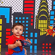 Thiago Armando is registered to the contest to win money with this photo: blue_shorts, building, child, cityscape, colorful_background, comic_style, curly_hair, cute, expression, floor, footwear, indoor, looking_away, playful, portrait, red_shirt, shoe, sitting, toddler, young_child