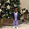 Joab is registered to the contest to win money with this photo: child, toddler, christmas_tree, ornaments, lights, lantern, holiday, decorations, poinsettia, purple_clothing, jacket, white_shoes, indoor, floor, festive, portrait, person, standing, young_child, celebration