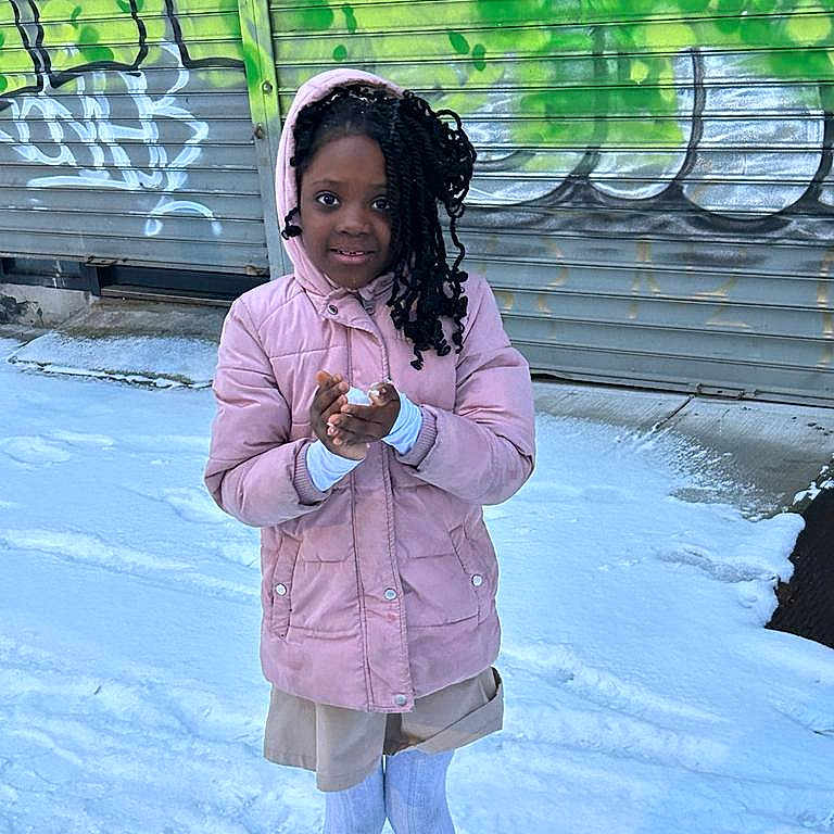 Jemima is registered to the contest to win money with this photo: child, girl, pink_coat, snow, winter, graffiti, urban, street, sidewalk, tights, boots, hood, clothing, outdoor, cold, building, metal_shutter, smiling, standing, portrait