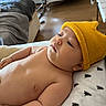 baby, baby_furniture, blanket, child, chubby, cozy, cute, hat, home, indoor, infant, lying_down, patterned_pillow, pillow, portrait, relaxed, room, skin, soft_lighting, yellow_hat