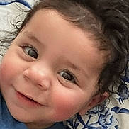 Tristan joined the competition — help win amazing prizes! baby, infant, smiling, curly_hair, dark_hair, cheeks, eyes, hand, blue_clothing, blanket, floral_pattern, portrait, close_up, bed, pillow, cute, skin, happy, playful, lying_down