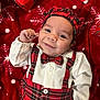 baby, infant, smiling, plaid_outfit, bow_tie, bonnet, red_blanket, christmas_gift, wrapping_paper, snowman, festive, holiday, portrait, lying_down, hand, eyes, cheek, button, fabric, cute