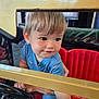 toddler, child, blue_clothing, red_seat, yellow_bars, steering_wheel, indoor, curious, playful, short_hair, face, hands, seat, toy_vehicle, person, smile, young_child, casual_clothing, light_hair, candid