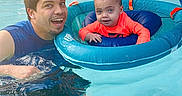 Zakiah is registered to the contest to win money with this photo: adult, baby, child, swimming_pool, pool_float, inflatable_float, water, outdoor, summer, family, father, parent, smiling, portrait, wet_hair, orange_shirt, blue_water, backyard, poolside, recreation