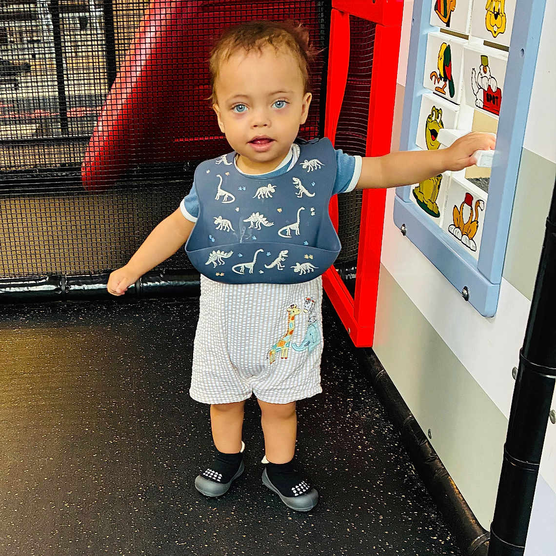 Zakiah is registered to the contest to win money with this photo: child, toddler, standing, dinosaur_shirt, shorts, shoes, smiling, blue_eyes, portrait, play_area, indoor, playground, red_slide, cartoon_panels, colorful, toy, activity_center, floor, play_structure, exploring