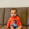 Zakiah is registered to the contest to win money with this photo: baby, child, toddler, sitting, couch, sofa, portrait, indoor, home, orange_shirt, dinosaur_shirt, barefoot, smiling, looking_at_camera, short_hair, facial_expression, furniture, wall_art, framed_picture, cozy