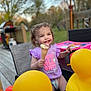 toddler, child, ice_cream, cone, purple_shirt, outdoor, patio, yellow_balloon, rubber_duck, smiling, curly_hair, happy, summer, playful, table, chair, nature, grass, daylight, fun
