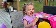 Olivia is registered to the contest to win money with this photo: chair, child, deck, fun, grass, happy, ice_cream, laughing, outdoor, person, plastic_toy, play_table, purple_shirt, rubber_duck, smiling, summer, table, toddler, trees, yellow