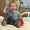 child, toddler, hospital_bed, red_cast, curly_hair, barefoot, gray_tshirt, camouflage_pants, plastic_bottle, smiling, medical_room, sink, light_switch, cabinet, playful, indoor, person, sitting, happy, young_child