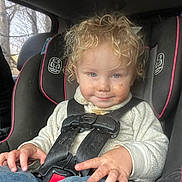 Adam is registered to the contest to win money with this photo: toddler, child, car_seat, curly_hair, smiling, face_smudge, hands, clothing, sweater, jeans, seatbelt, vehicle_interior, window, trees, daylight, candid, portrait, safety, cute, messy