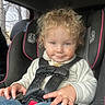 toddler, child, car_seat, curly_hair, smiling, face_smudge, hands, clothing, sweater, jeans, seatbelt, vehicle_interior, window, trees, daylight, candid, portrait, safety, cute, messy