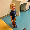 Adam is registered to the contest to win money with this photo: toddler, child, curly_hair, blue_shirt, brown_pants, blue_shoes, floor, yellow_floor, blue_floor, sink, cabinet, indoor, person, curious_expression, standing, footwear, boot, casual, room, wall