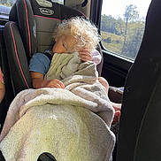 Adam is registered to the contest to win money with this photo: blanket, car_interior, car_seat, casual_clothing, child, comfort, cozy, curly_hair, daytime, nap, relaxing, road_trip, seat, seatbelt, sleepy, sunlight, toddler, travel, window, young_child