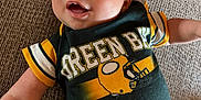 Cooper is registered to the contest to win money with this photo: baby, blanket, child, clothing, couch, cute, eyes, fabric, face, green_bay_packers, happy, head, indoors, infant, laying_down, portrait, skin, sleeves, smiling, sportswear