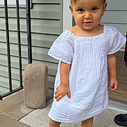Daphne is registered to the contest to win money with this photo: toddler, child, white_dress, pink_sandals, bows, porch, curious, hair_accessories, footwear, outdoor, sidewalk, railing, bracelet, person, young_child, standing, smiling, cute, fashion, summer