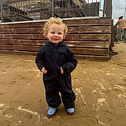Caleb-Jack joined the competition — help win amazing prizes! child, toddler, curly_hair, smiling, black_jumpsuit, blue_boots, sand, beach, fence, wood, outdoor, cloudy_sky, person, happy, standing, footprints, casual_clothing, daytime, young_child, playful
