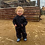 child, toddler, curly_hair, smiling, black_jumpsuit, blue_boots, sand, beach, fence, wood, outdoor, cloudy_sky, person, happy, standing, footprints, casual_clothing, daytime, young_child, playful