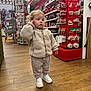 toddler, child, curly_hair, beige_jacket, white_sneakers, pants, wood_floor, christmas_decorations, ornaments, store_aisle, holiday, red_display, shopping, indoor, curious, person, festive, winter_clothing, retail, young_child