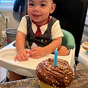 Jayden joined the competition — help win amazing prizes! toddler, child, high_chair, cupcake, birthday_candle, chocolate_frosting, sprinkles, smiling, indoor, kitchen, table, celebration, person, food, dessert, happy, curly_hair, short_sleeve_shirt, plastic_seat, party