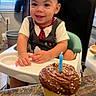 toddler, child, high_chair, cupcake, birthday_candle, chocolate_frosting, sprinkles, smiling, indoor, kitchen, table, celebration, person, food, dessert, happy, curly_hair, short_sleeve_shirt, plastic_seat, party
