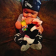 Armani is registered to the contest to win money with this photo: baby, black_clothing, chewing, child, colorful_lighting, couch, cozy, feet, hands, hat, indoor, infant, photograph, plush_toy, portrait, puma, sitting, socks, sportswear, toy