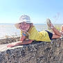 Louyana participe au concours pour gagner de l'argent avec cette photo : child, hat, yellow_shirt, stone_wall, outdoor, sunny, lake, water, blue_sky, plants, sandals, relaxing, smiling, casual, nature, summer, bracelets, legs, portrait, person