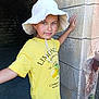 child, hat, yellow_shirt, lemon_graphics, stone_wall, archway, earrings, summer, outdoor, portrait, smile, casual_clothing, young_girl, sun_hat, texture, shadow, posing, happy, person, daylight