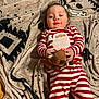 baby, child, pajamas, striped_clothing, plush_toy, blanket, floor, indoor, cute, holiday_theme, face, person, infant, red, white, patterned_blanket, toy, holding, lying_down, curly_hair