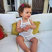 Maëna participe au concours pour gagner de l'argent avec cette photo : child, toddler, curly_hair, hair_clip, white_couch, cushion, knitted_romper, striped_clothing, barefoot, indoors, window, door, wooden_shutters, smiling, portrait, sitting, cute, expression, skin, furniture