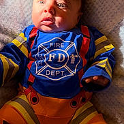 Maverick is registered to the contest to win money with this photo: baby, child, firefighter_costume, blue_shirt, orange_pants, infant, cute, soft_blanket, portrait, young_child, costume, bright_colors, face, hand, expression, laying_down, indoors, clothing, person, baby_face