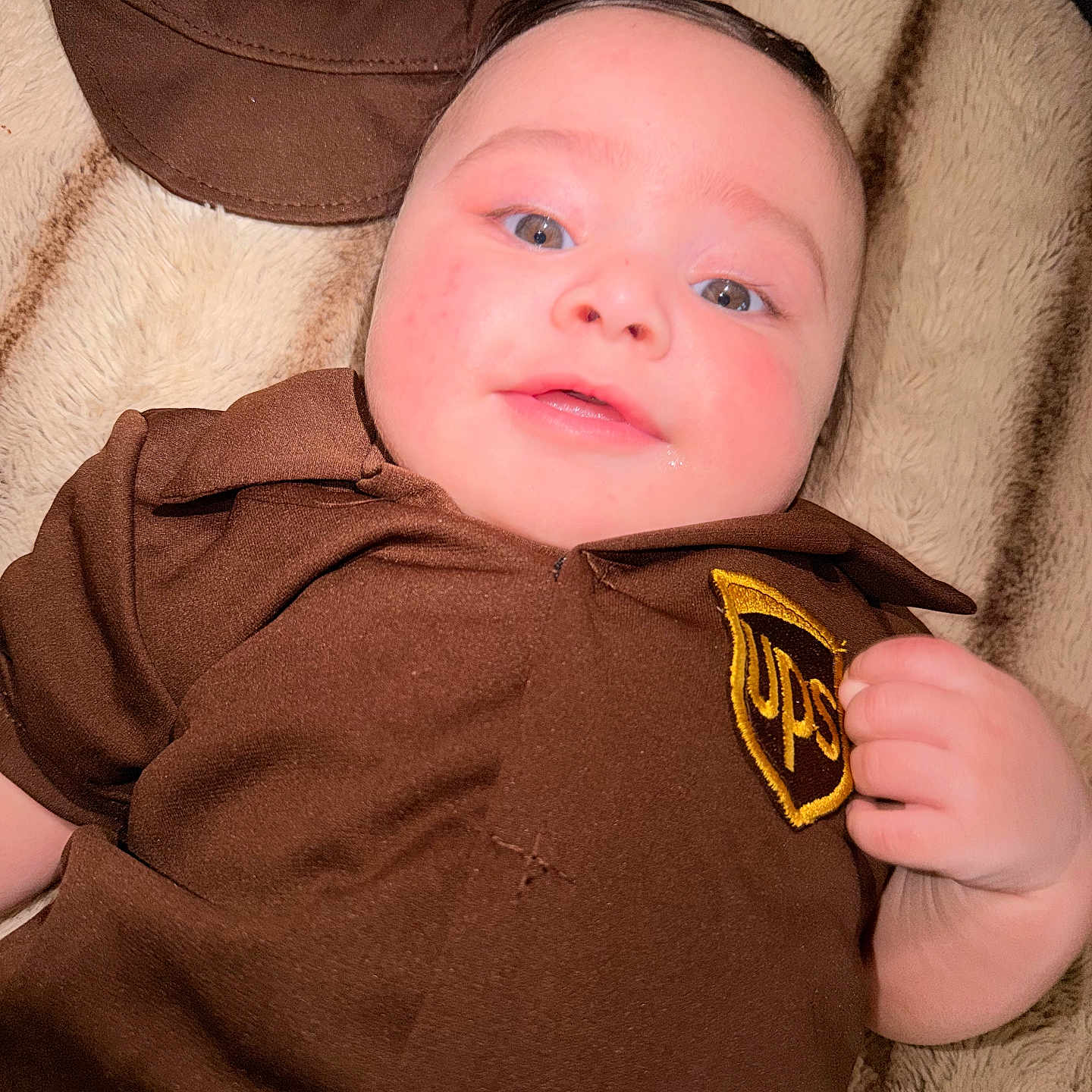 Maverick joined the competition — help win amazing prizes! baby, infant, child, face, person, clothing, uniform, brown, hat, logo, ups, blanket, lying_down, cute, drool, expression, portrait, indoors, hand, closeup