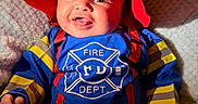 Maverick joined the competition — help win amazing prizes! baby, child, costume, firefighter, red_hat, blue_shirt, orange_pants, smiling, tongue_out, cute, happy, lying_down, blanket, soft_texture, indoor, portrait, person, face, hand, colorful_clothing