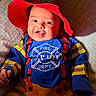 baby, child, costume, firefighter, red_hat, blue_shirt, orange_pants, smiling, tongue_out, cute, happy, lying_down, blanket, soft_texture, indoor, portrait, person, face, hand, colorful_clothing
