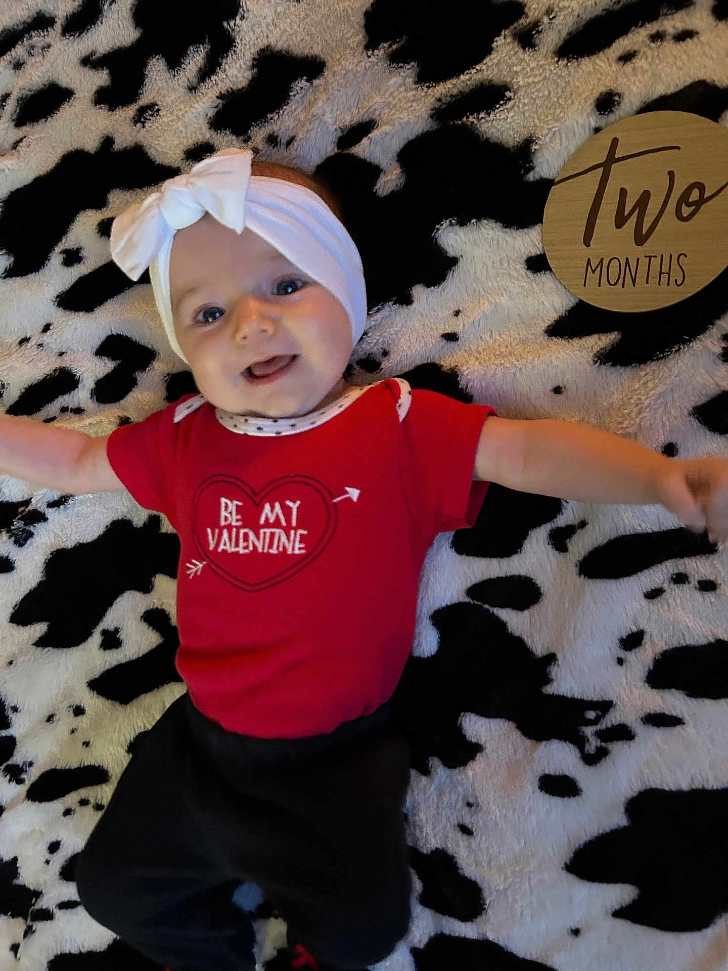 Alora is registered to the contest to win money with this photo: baby, infant, headband, bow, onesie, valentine, red_clothing, smile, tongue_out, blanket, cow_print, milestone_sign, wooden_sign, two_months, lying_down, arms_outstretched, adorable, portrait, indoor, soft_texture