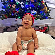 Amor is registered to the contest to win money with this photo: baby, smiling, red_beanie, red_slippers, diaper, blanket, christmas_tree, christmas_lights, plush_toys, toy_train, floor, indoor, happy, child, holiday, festive, decorations, candid, cute, joyful