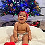 baby, smiling, red_beanie, red_slippers, diaper, blanket, christmas_tree, christmas_lights, plush_toys, toy_train, floor, indoor, happy, child, holiday, festive, decorations, candid, cute, joyful