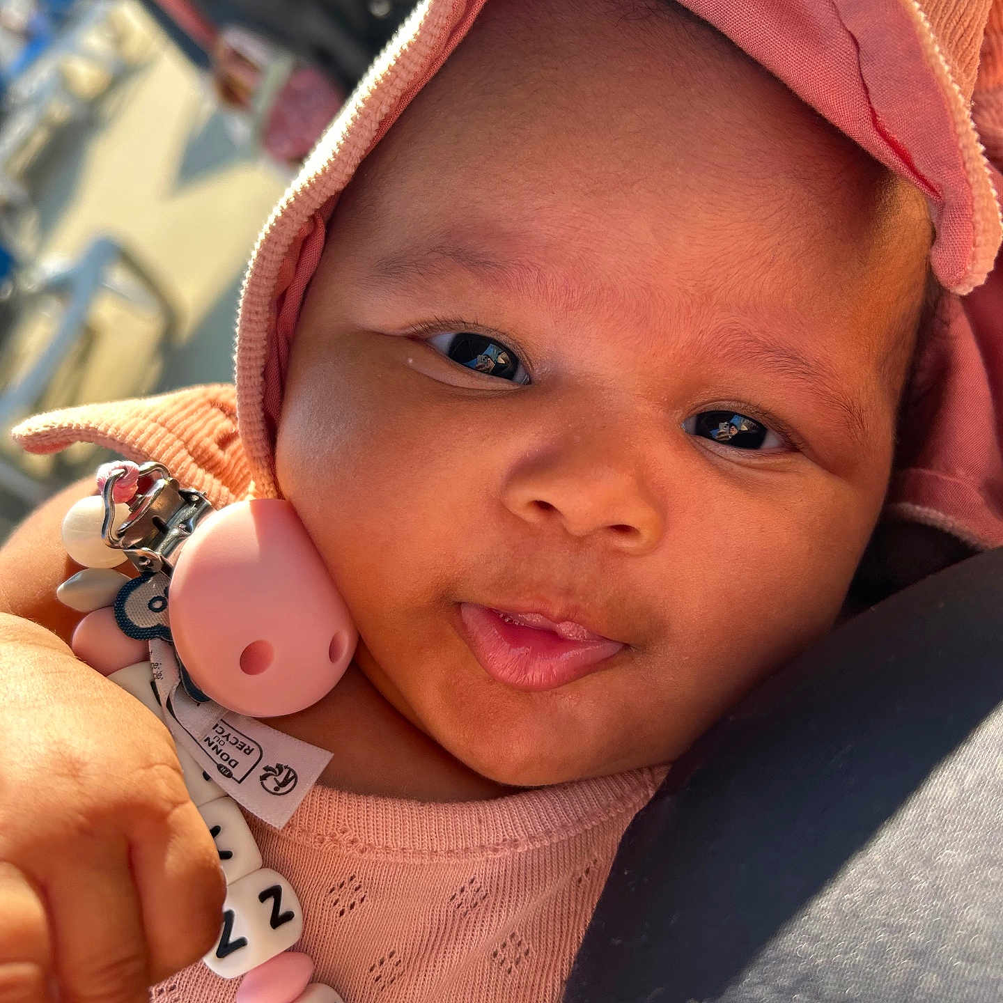 Loa Nawi participe au concours pour gagner de l'argent avec cette photo : baby, beads, child, closeup, clothing, cute, expression, face, hand, holding, hood, infant, outdoor, pacifier, person, pink, portrait, skin, warm_light, young_child