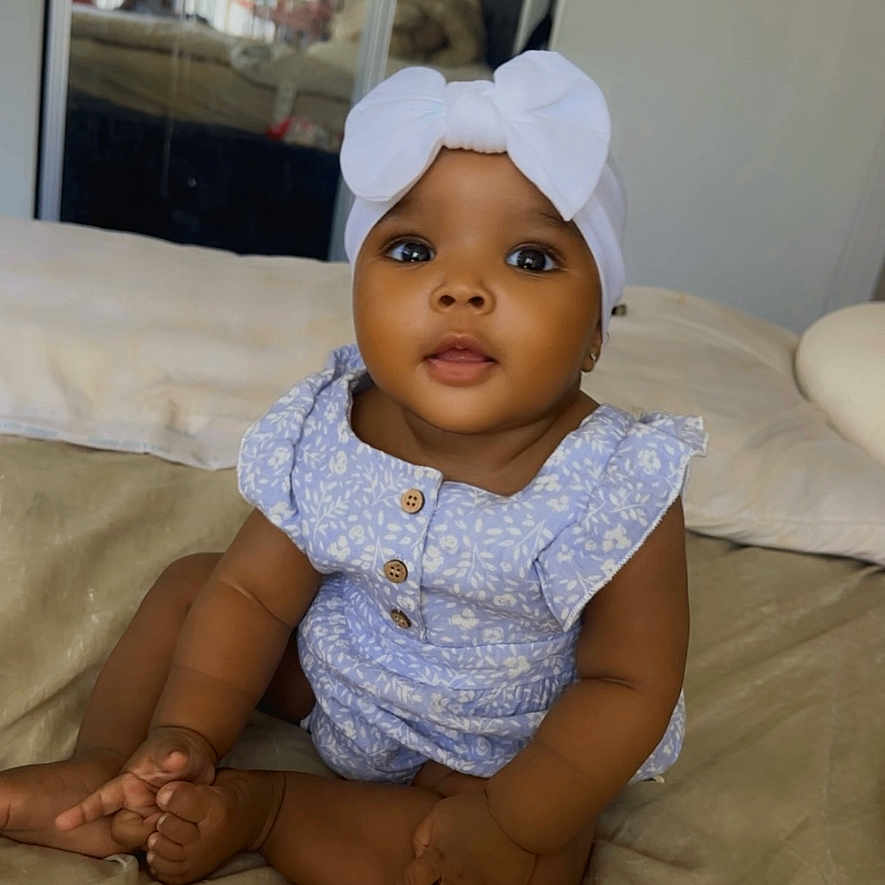 Germany is registered to the contest to win money with this photo: baby, child, infant, portrait, sitting, bed, bedroom, pillow, bow, headband, dress, floral_pattern, smiling, face, eyes, indoor, home, mirror, furniture, photography