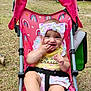 Eliza is registered to the contest to win money with this photo: baby, child, stroller, pink, rainbow_pattern, bow, yellow_clothing, outdoor, grass, smiling, cute, infant, playful, seatbelt, person, happy, sunlight, nature, headband, summer
